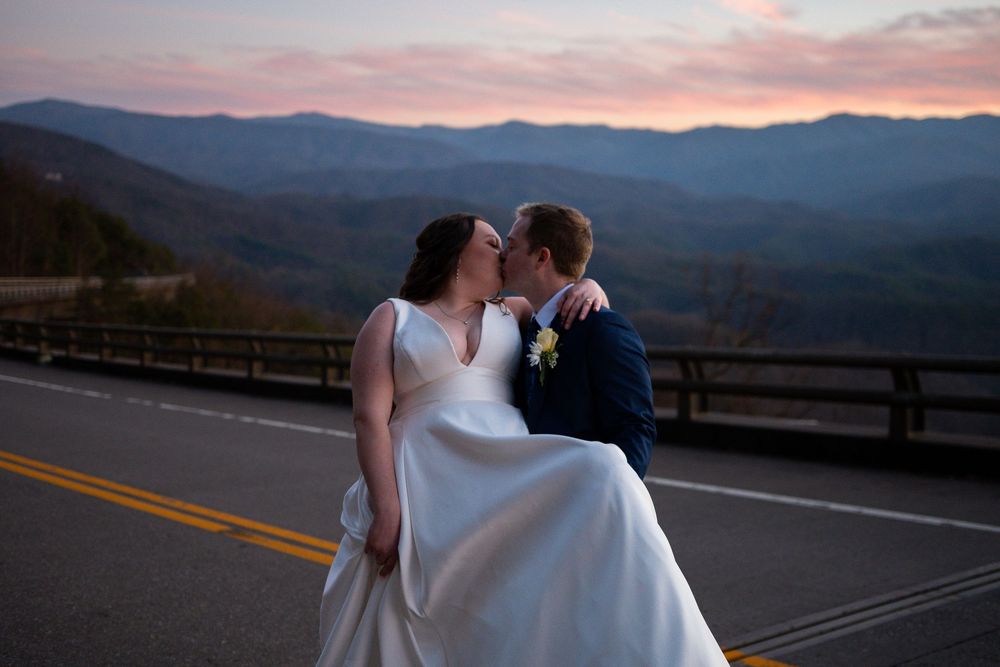 Wedding couple poses on scenic mountain overlook during a pink and purple sunset.