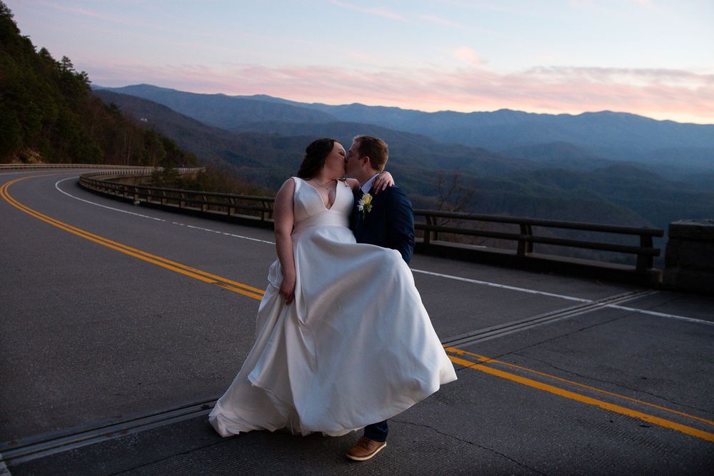 Couple in wedding attire dances on scenic mountain highway at sunset.