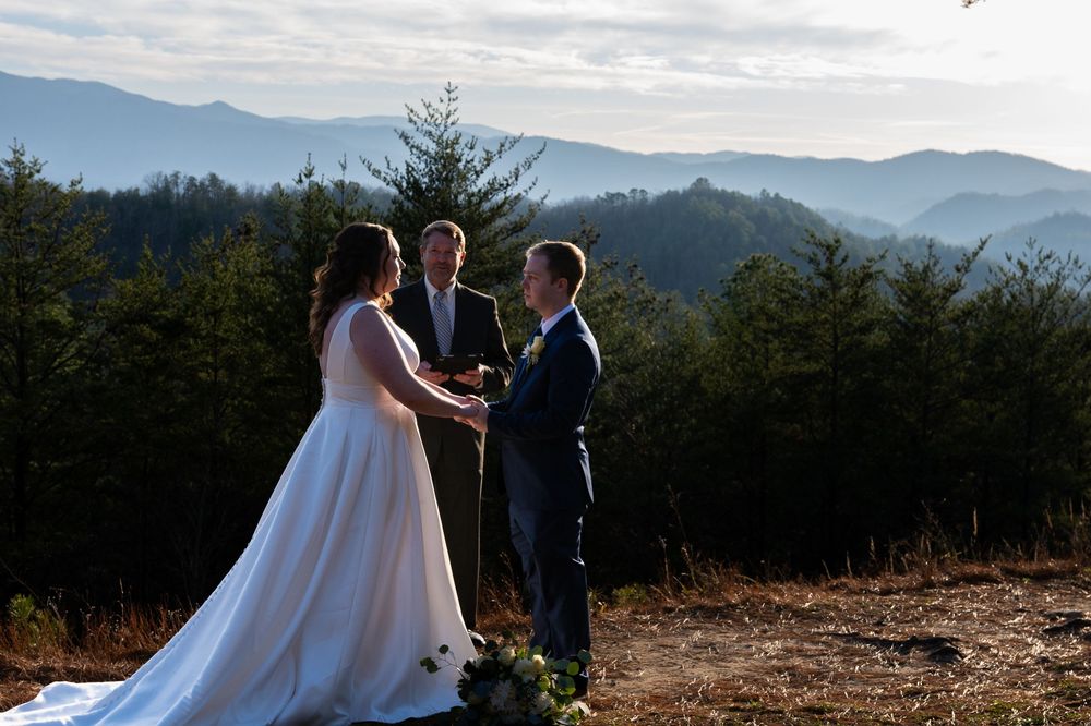 A wedding ceremony takes place on a mountaintop with evergreen trees and misty peaks in the background.