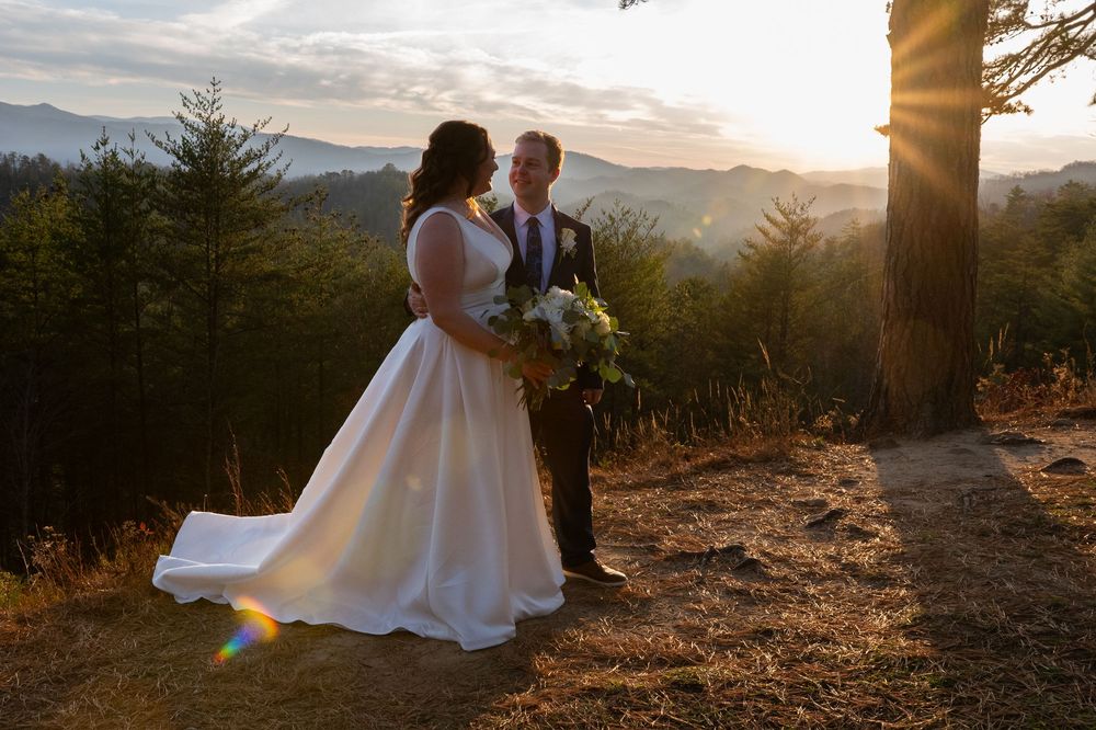 A couple shares a tender moment at sunset on a mountaintop with scenic mountain views in the background.