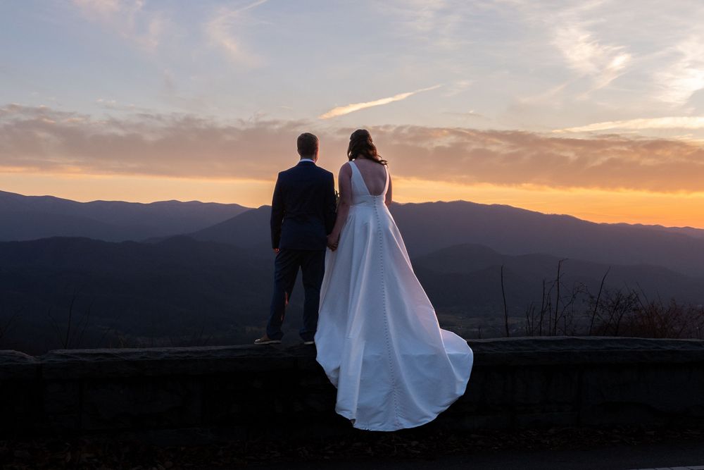 A couple stands silhouetted against a mountain sunset with a flowing white wedding dress.