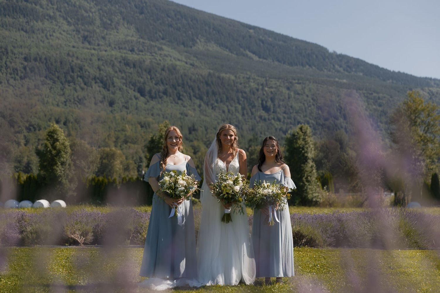 Bride and two bridesmaids in blue dresses hold bouquets in a lavender field with mountain backdrop.