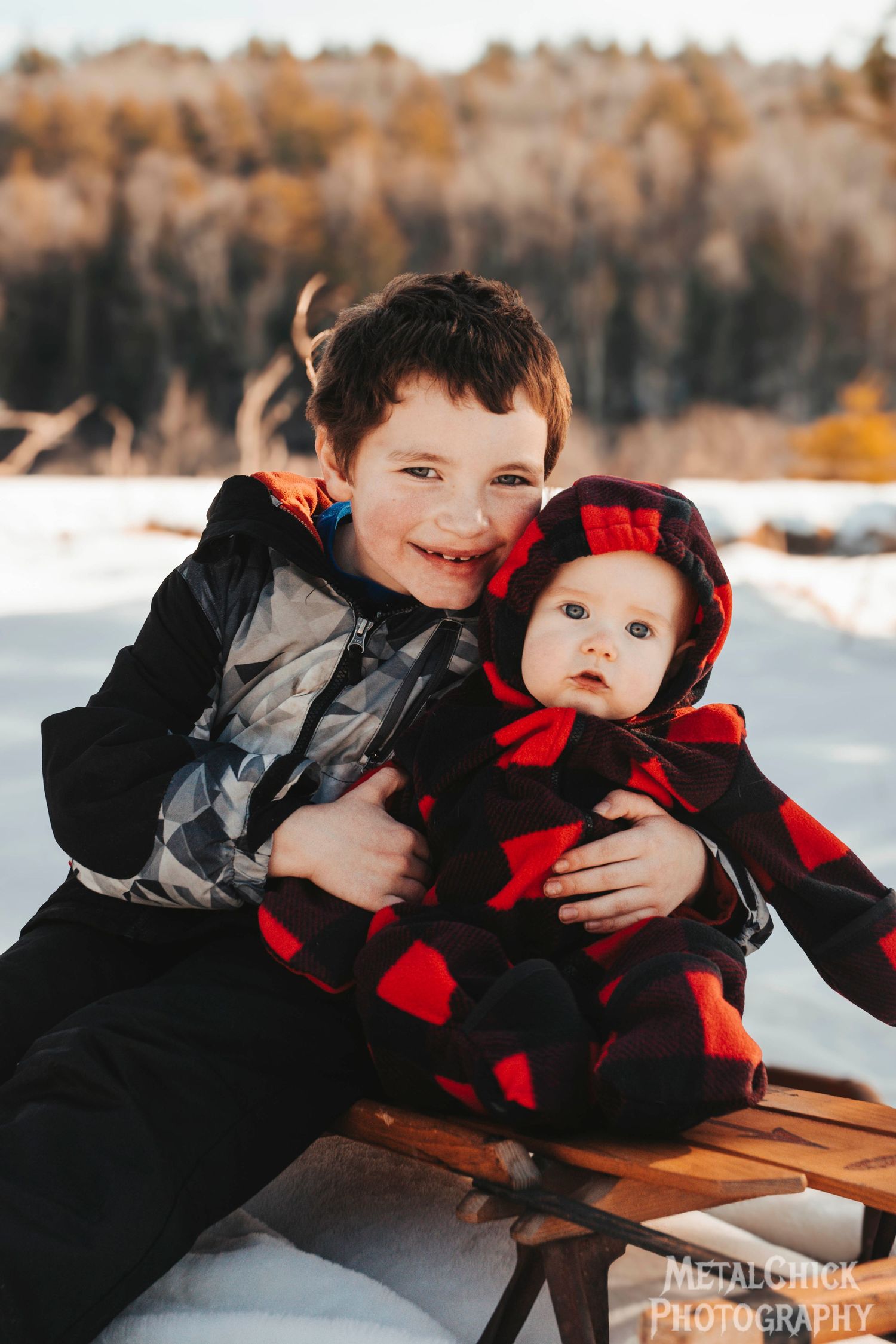 Children portrait by Northern Exposure featuring two brothers sitting on a runner sled in the middle of a snowy field with mountains.