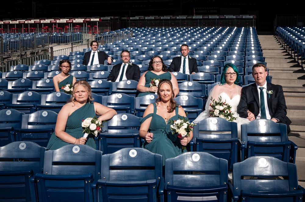Wedding party members in formal attire seated in rows of empty blue stadium seats at a sports venue.