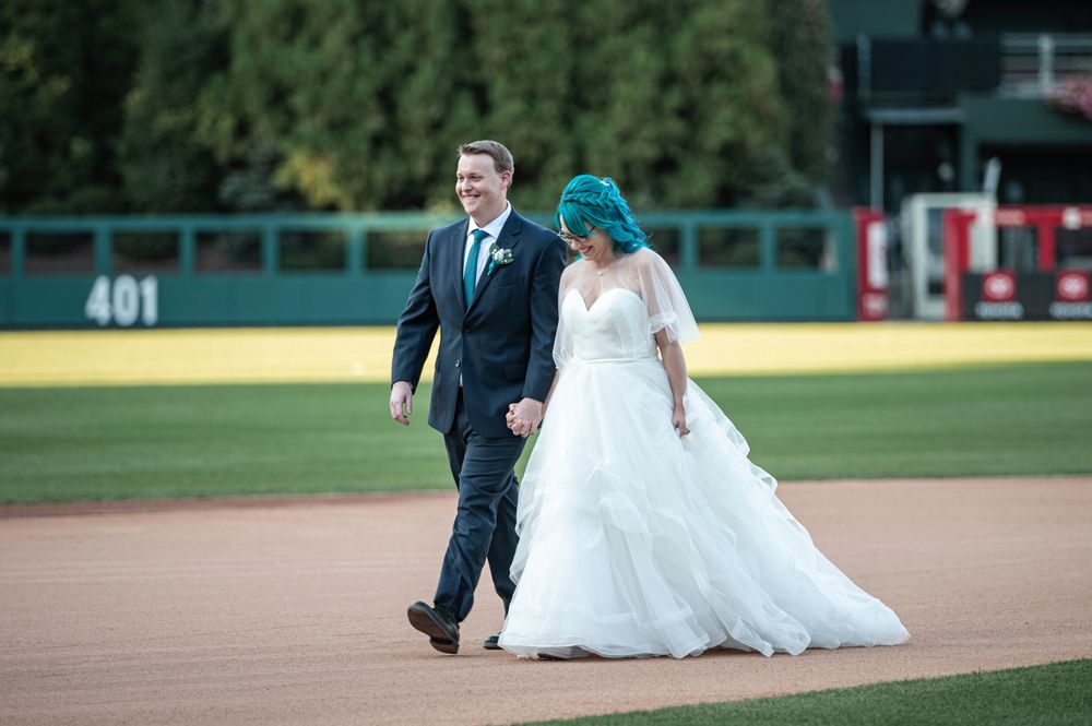 A couple walking together on a baseball field with the bride wearing a flowing white dress and groom in dark suit.