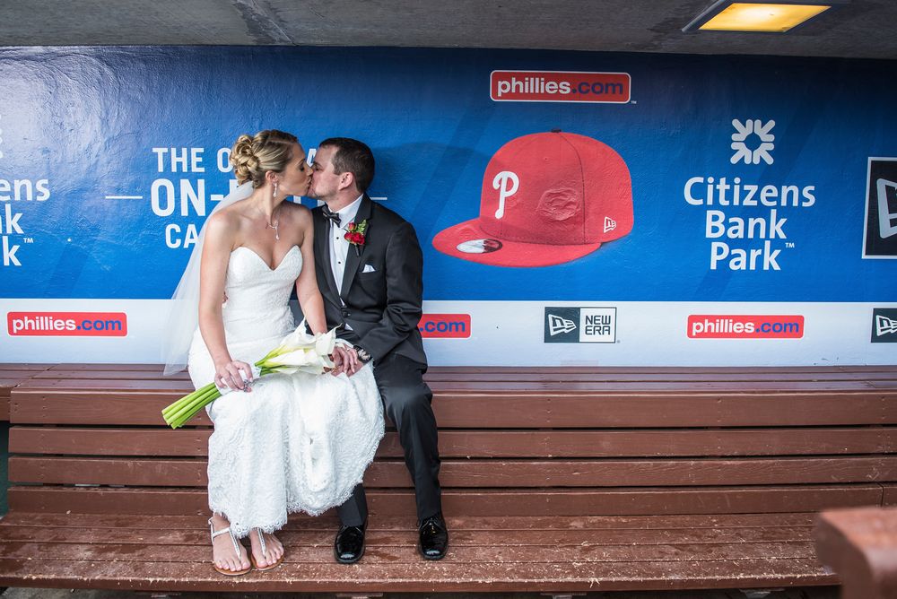 A wedding couple shares a romantic moment in a baseball dugout at Citizens Bank Park with Phillies decor in the background.