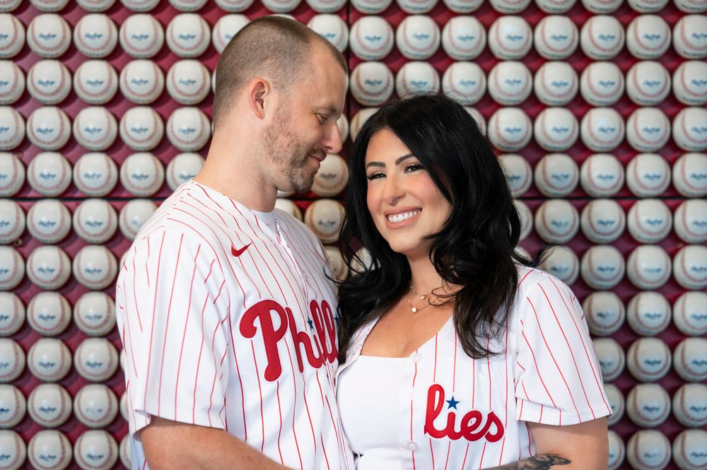 A couple wearing matching Philadelphia Phillies baseball jerseys smile at each other in front of a wall of baseballs.