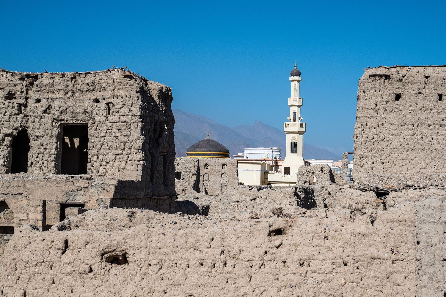 White minaret of a mosque rises above crumbling mud brick ruins in an ancient Middle Eastern village under blue sky.