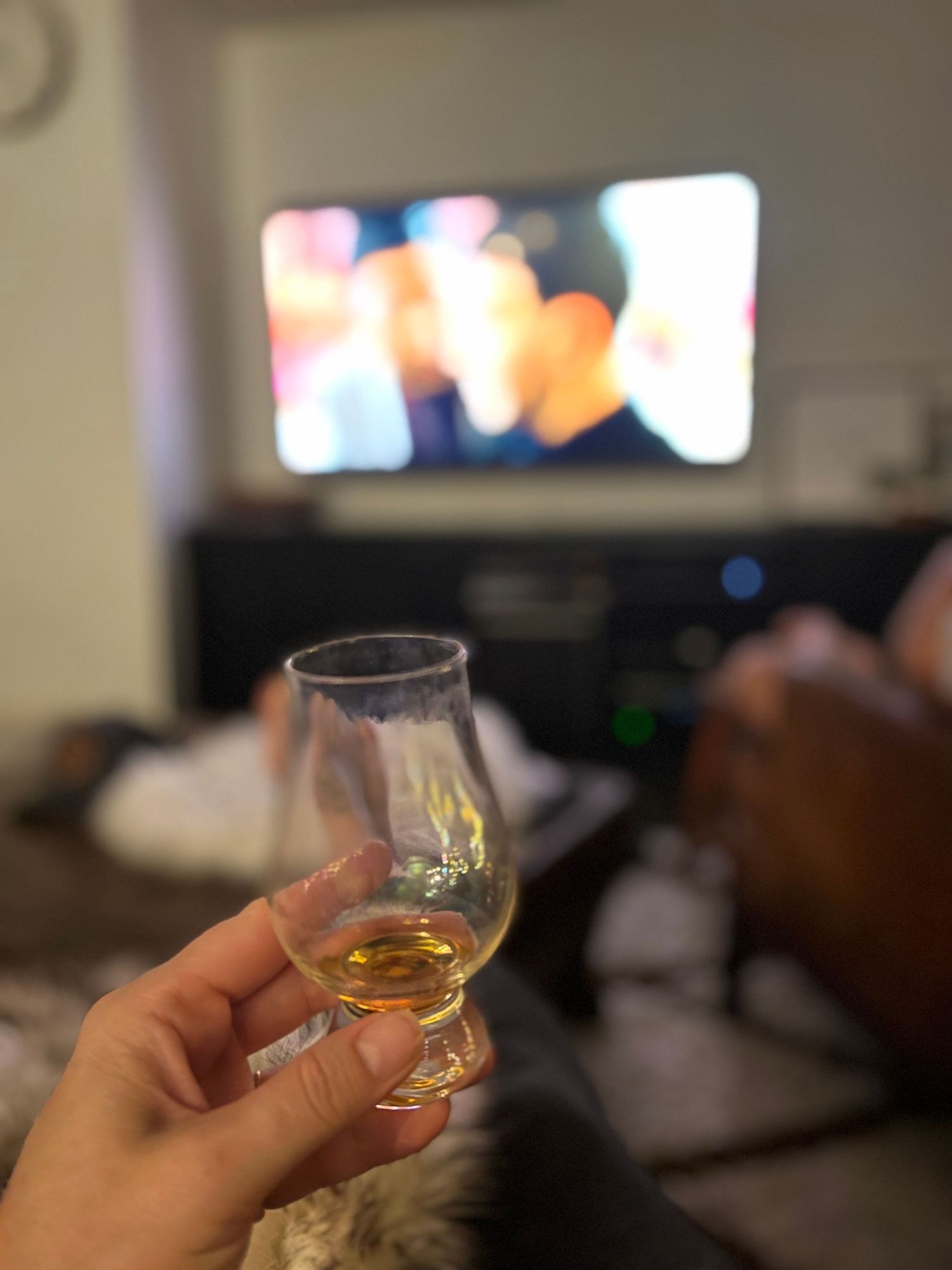 A hand holds a glencairn whiskey glass with golden spirit inside while watching TV in a dimly lit room.