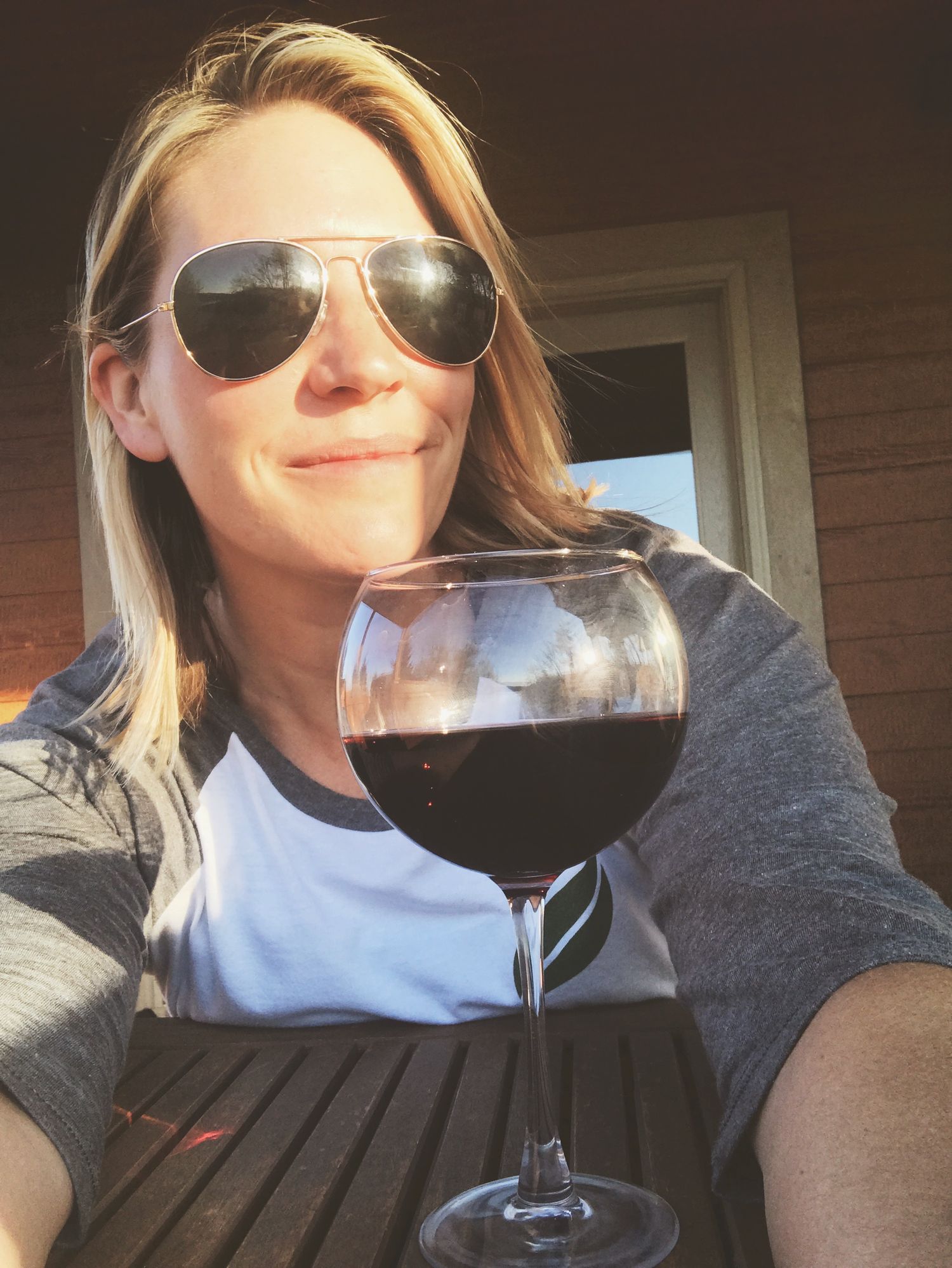 Person in sunglasses enjoys red wine on outdoor patio while relaxing in casual attire.
