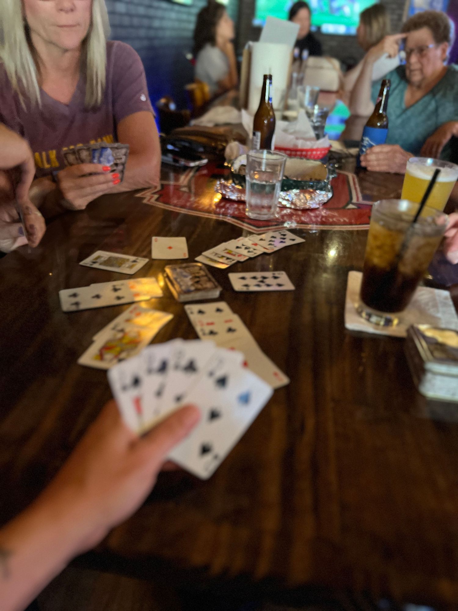 People playing cards at a wooden table with drinks and snacks visible in a dimly lit setting.