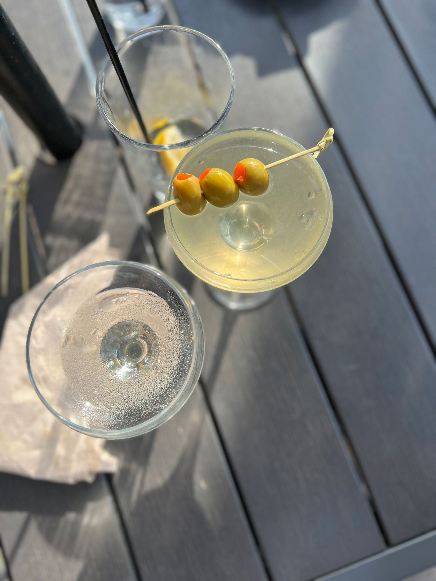 A martini glass filled with clear cocktail garnished with three green olives on a wooden deck in natural light.
