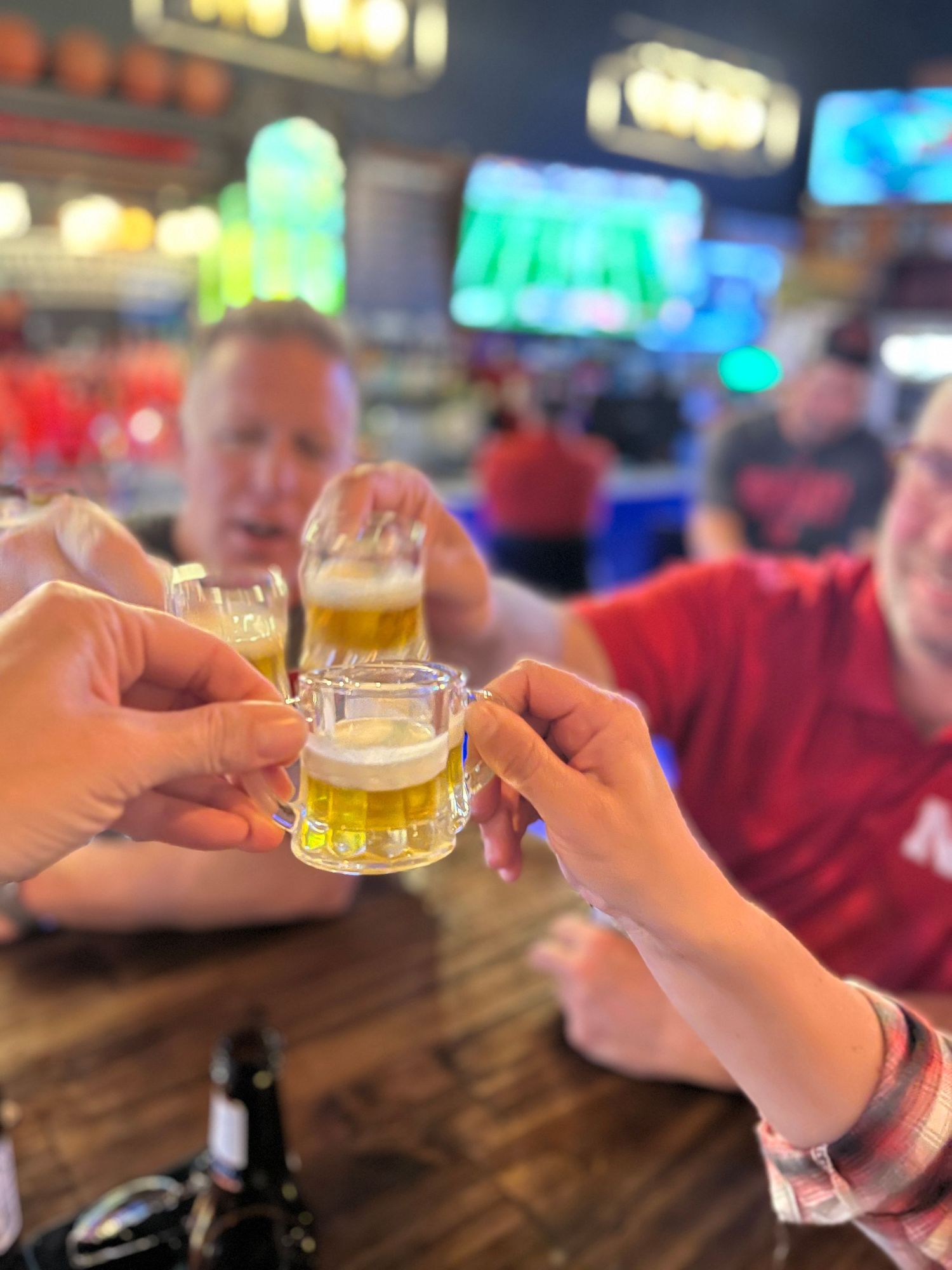 Friends clink small beer glasses in a toast at a dimly lit bar with colorful lights in the background.