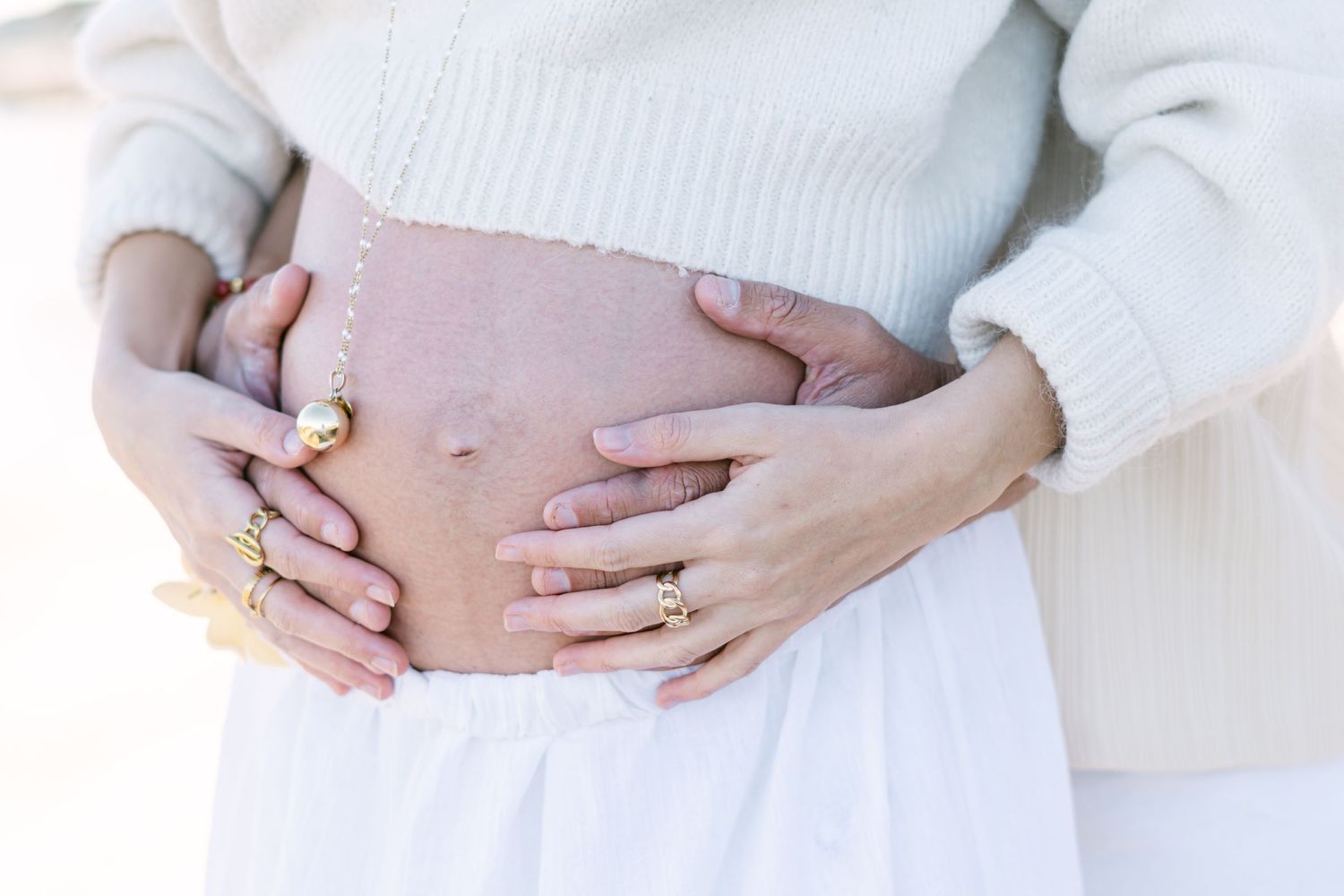 Hands gently cradling a pregnant belly while wearing a white sweater and skirt against a light background.