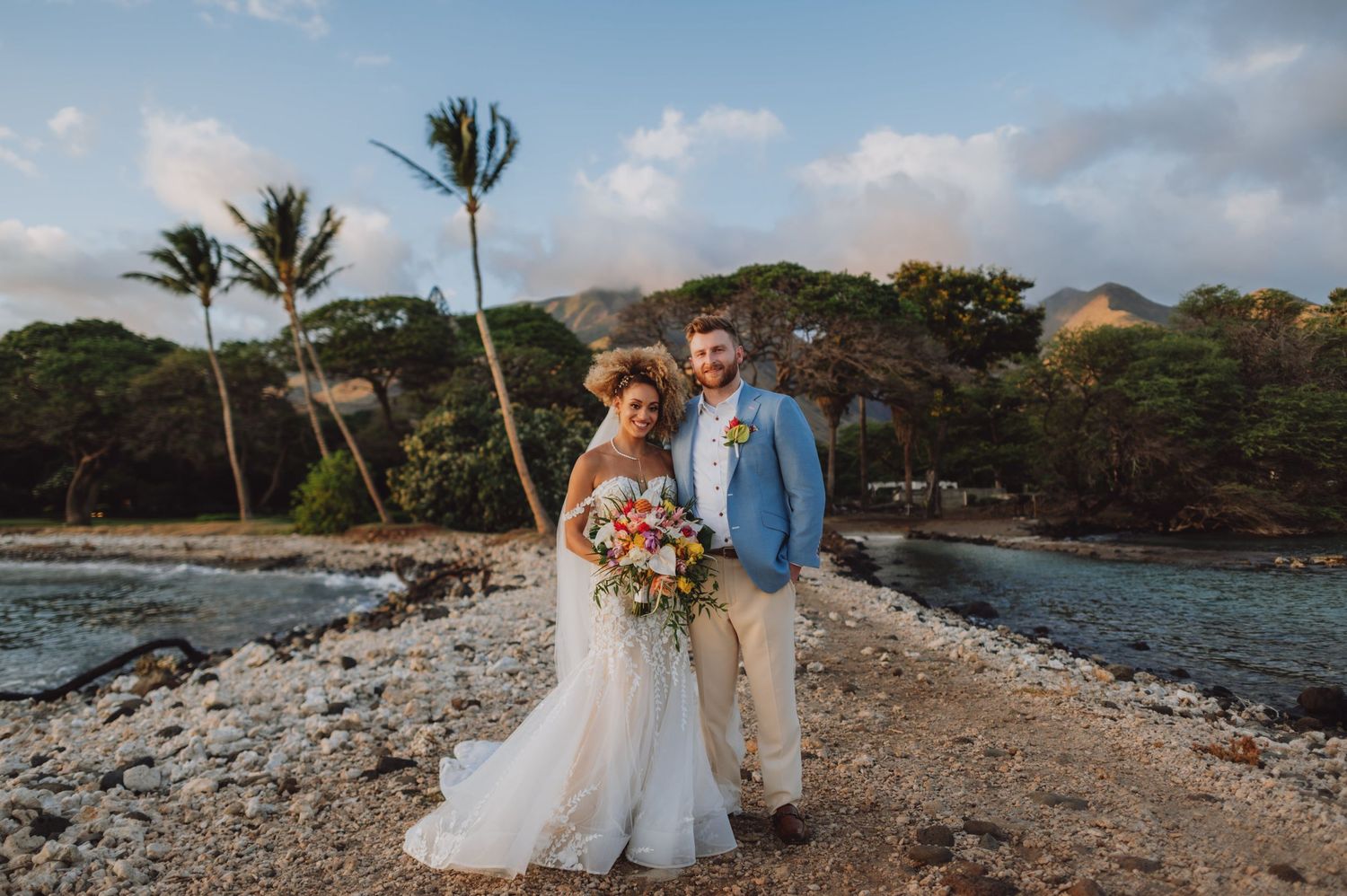 Wedding couple walking along rocky beach shore with palm trees and mountains in background.