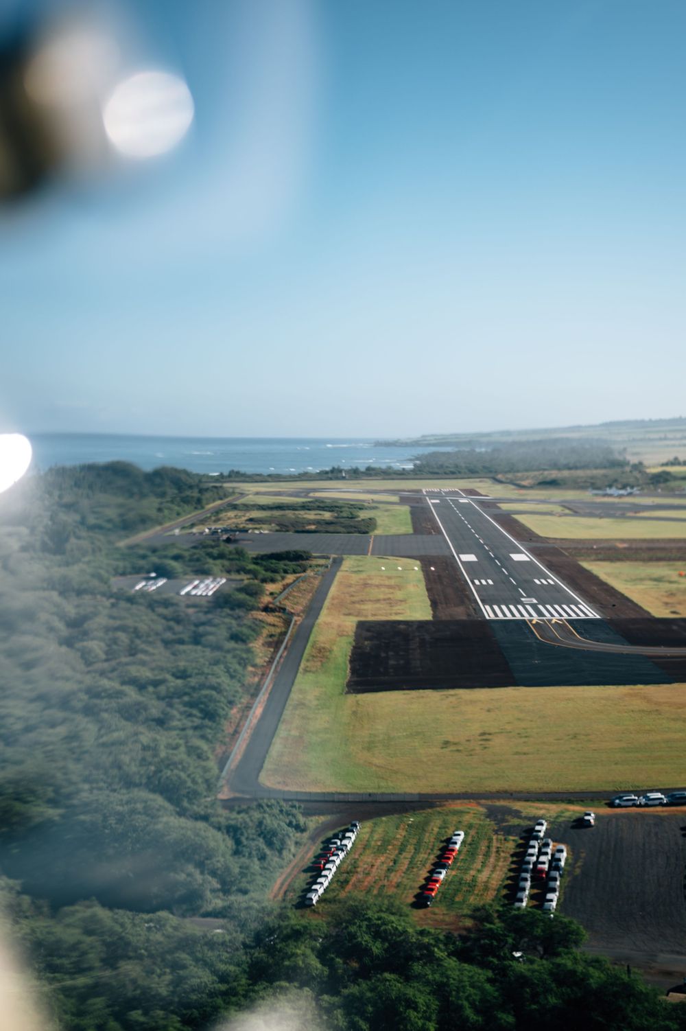 Bird's eye view of coastal airport runway with parking area and surrounding landscape.