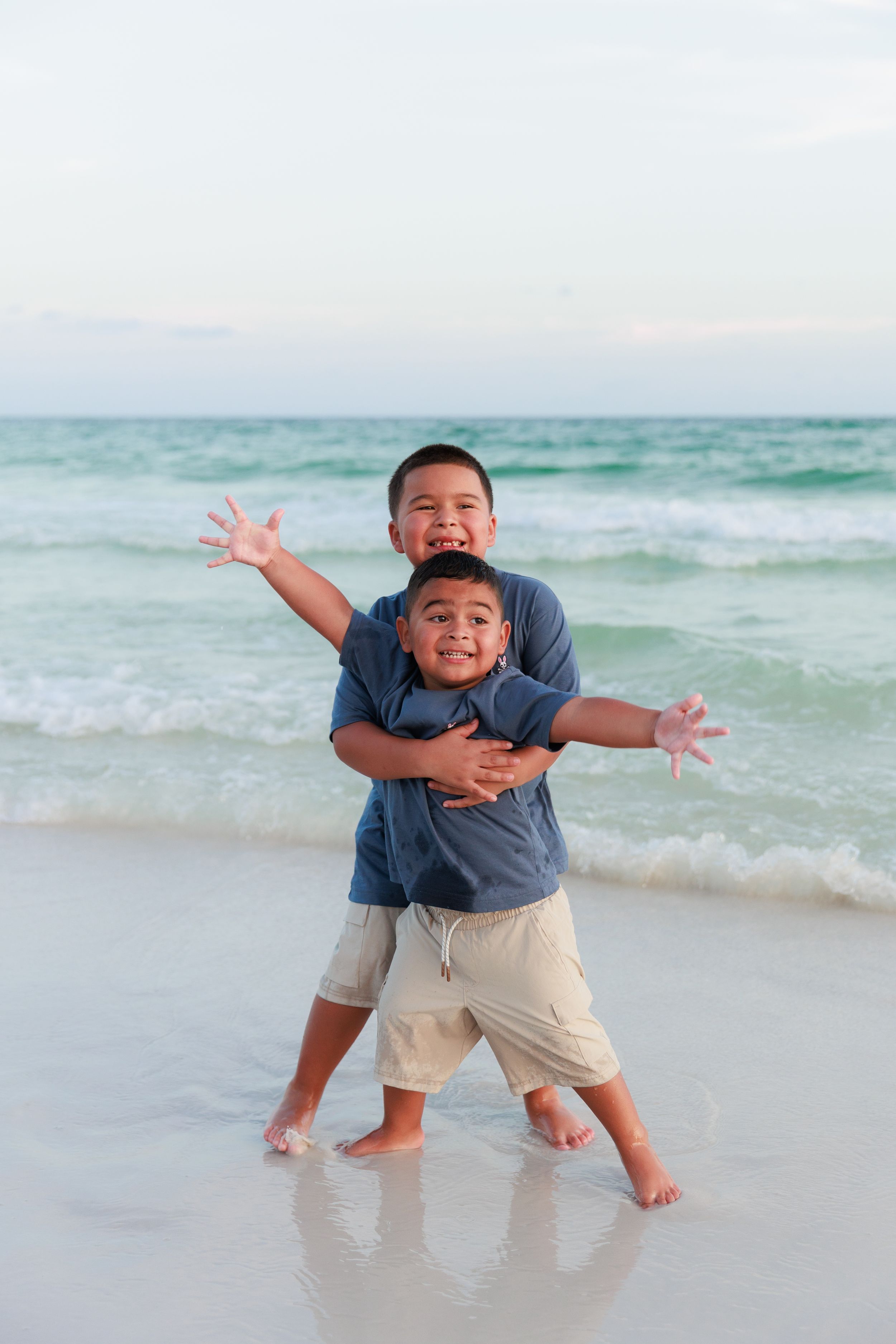 Playful child jumping and making expressive poses on a sunny beach with turquoise ocean waves in the background.