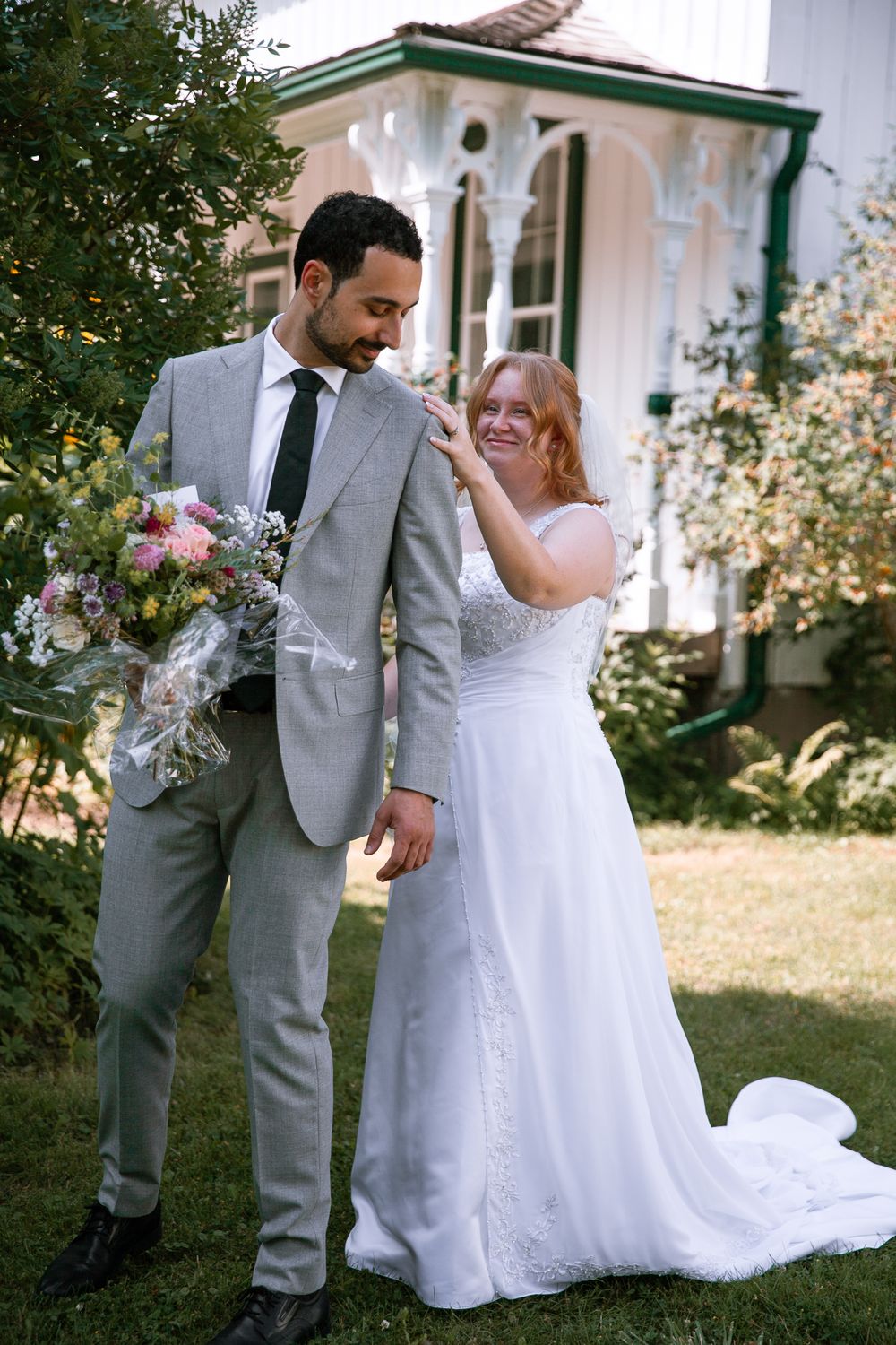 Bride in white gown laughs with groom in gray suit holding wildflower bouquet outside vintage home.