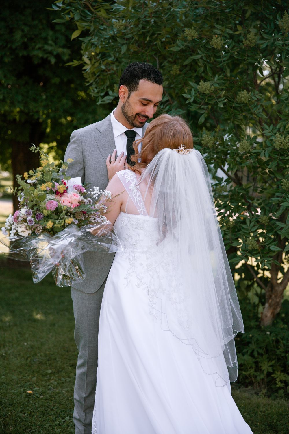 Bride and groom share intimate moment among lush green trees, bride holding colorful wildflower bouquet.