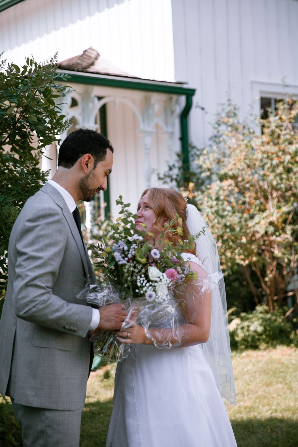Bride and groom gaze at each other holding wildflower bouquet outside white Victorian-style building.
