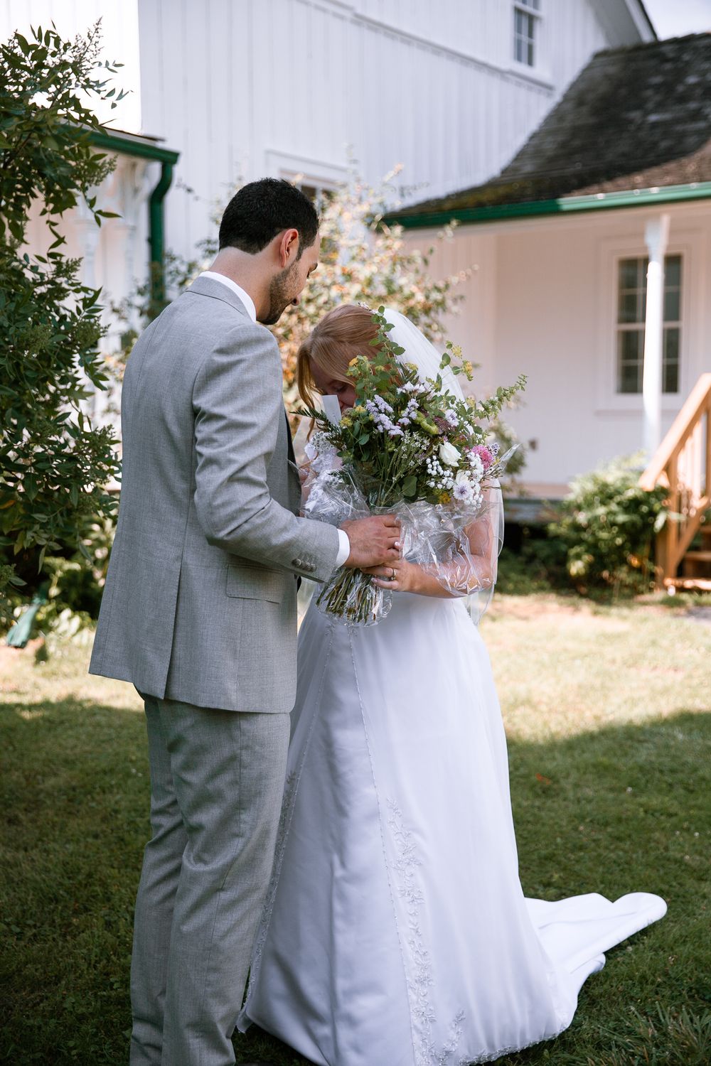 Bride and groom smell wildflower bouquet together outside white barn on sunny wedding day.