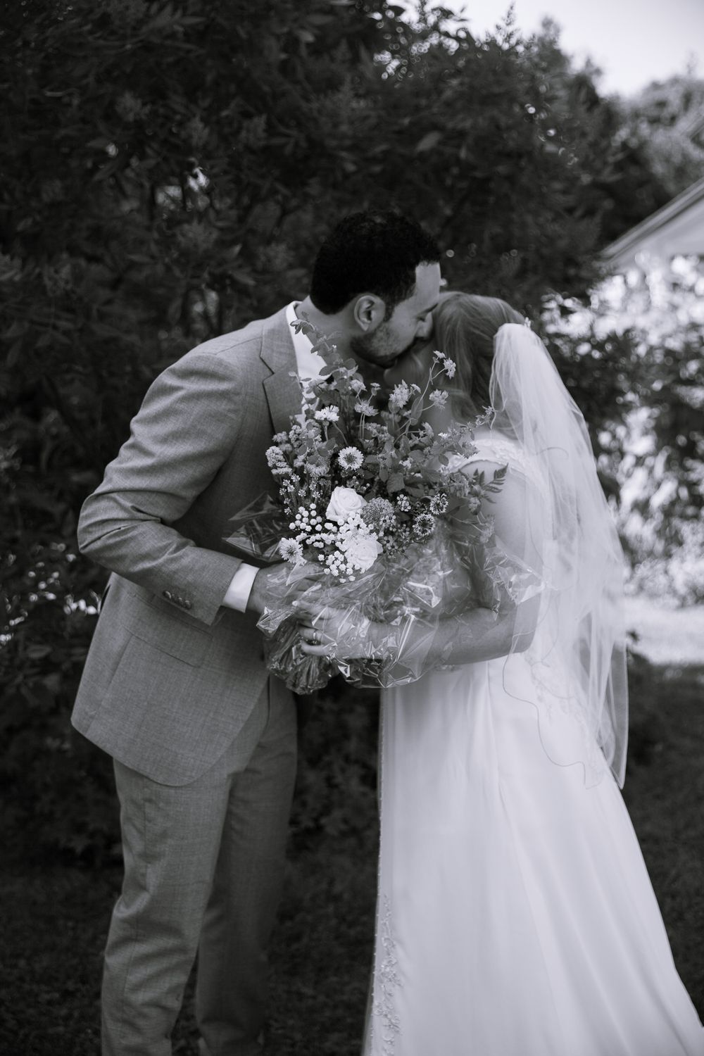 Black and white photo of bride and groom kissing behind lush wildflower wedding bouquet outdoors.