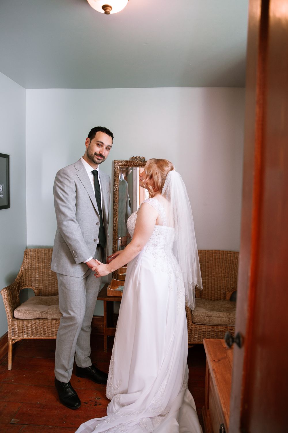 Smiling groom in gray suit holds bride's hands during indoor first look in rustic room.