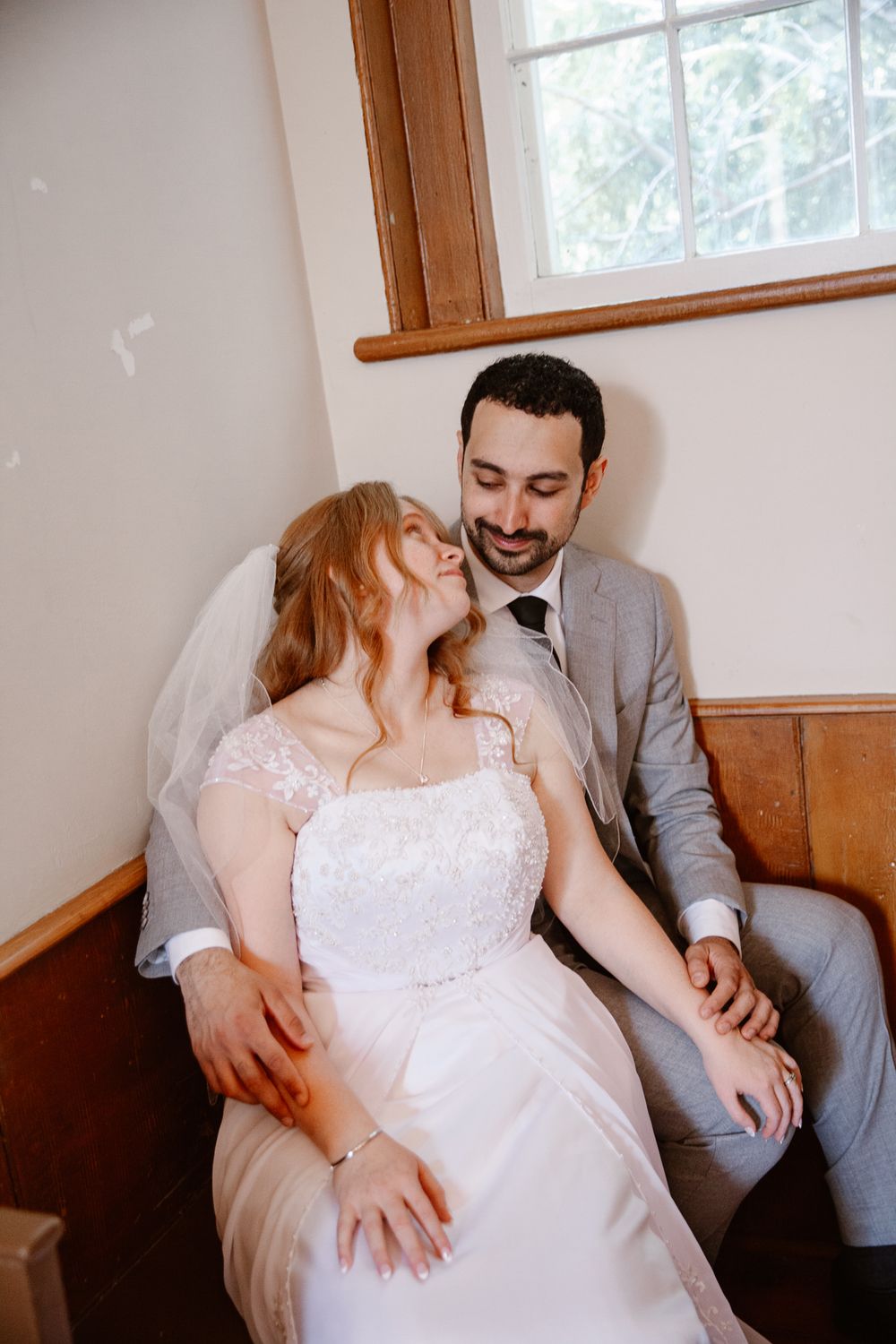 Bride in lace gown and groom in gray suit sit together on wooden bench by window.
