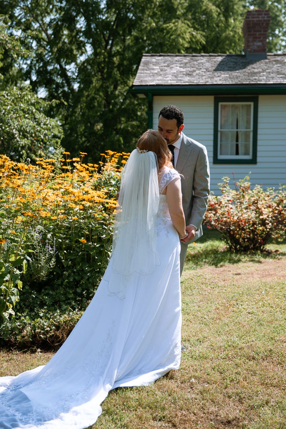 Groom kisses bride near vibrant yellow garden flowers outside charming blue cottage home.