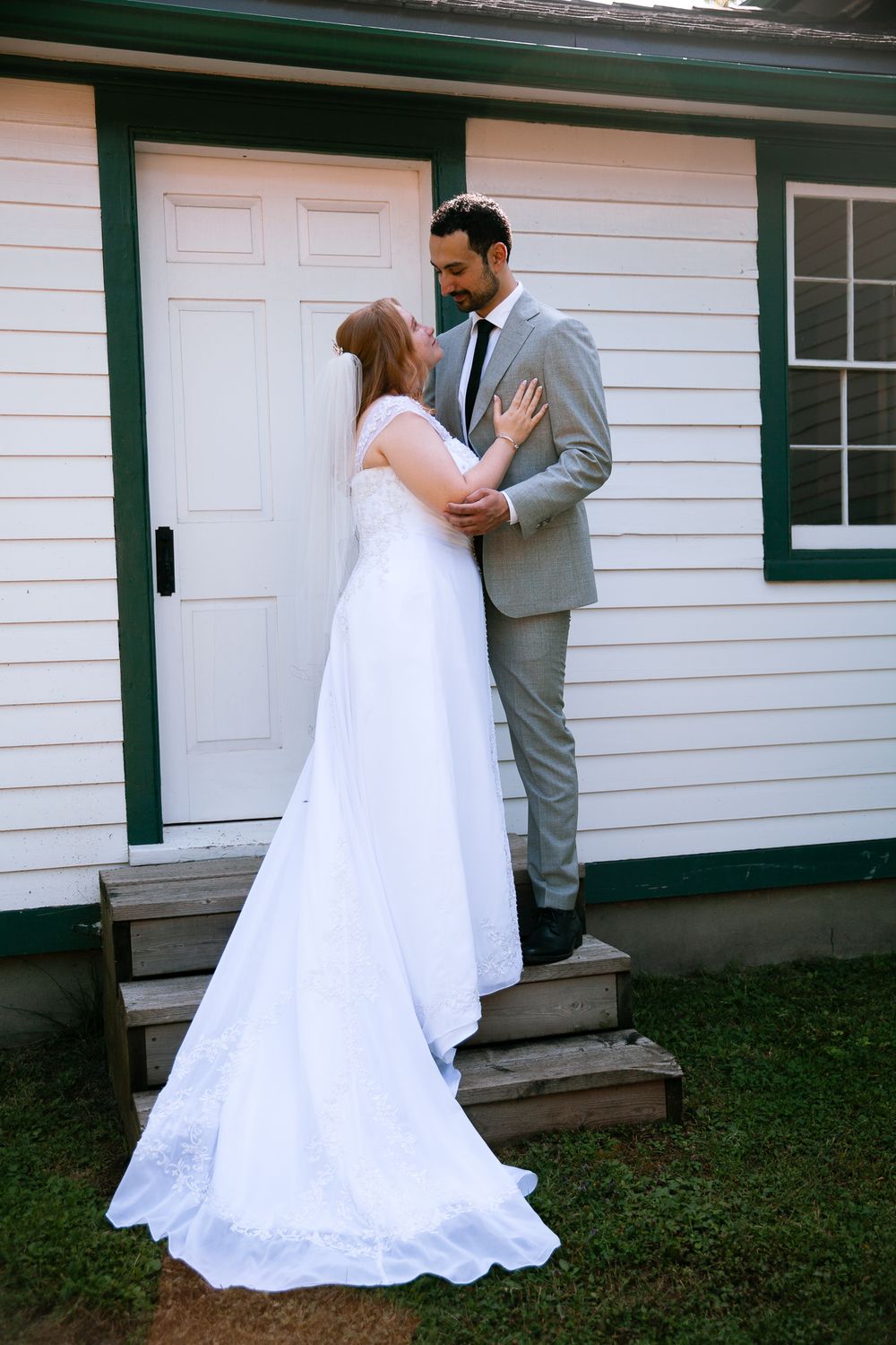 Bride and groom embrace on wooden steps of white building with green trim outdoors.