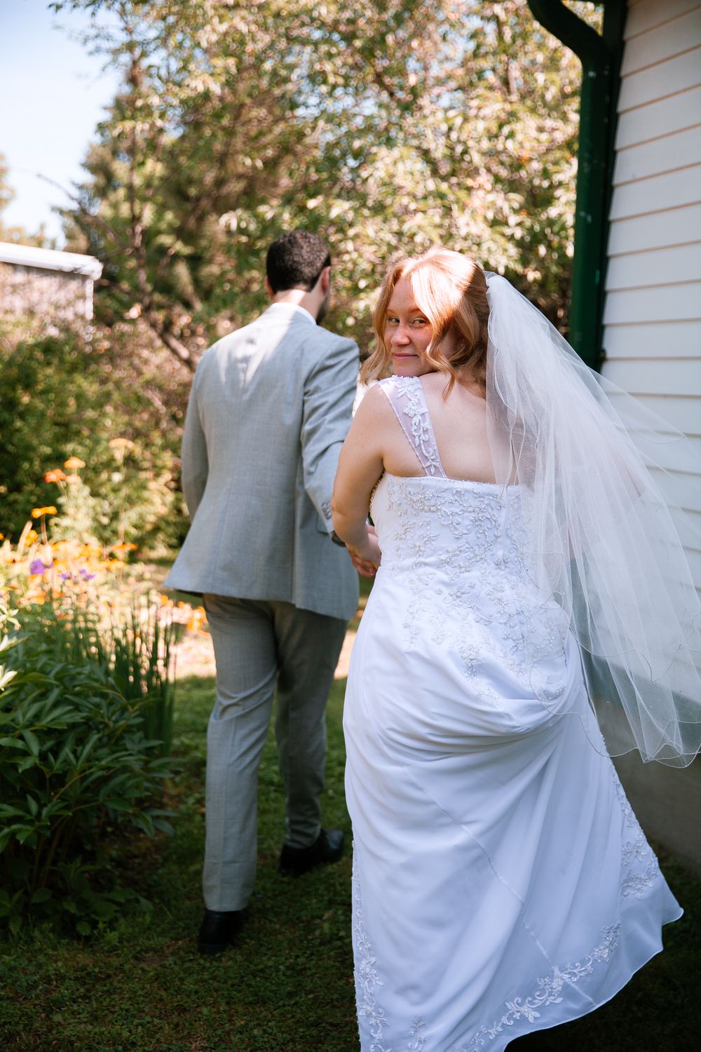 Bride in lace veil glances back smiling while walking with groom through garden path.