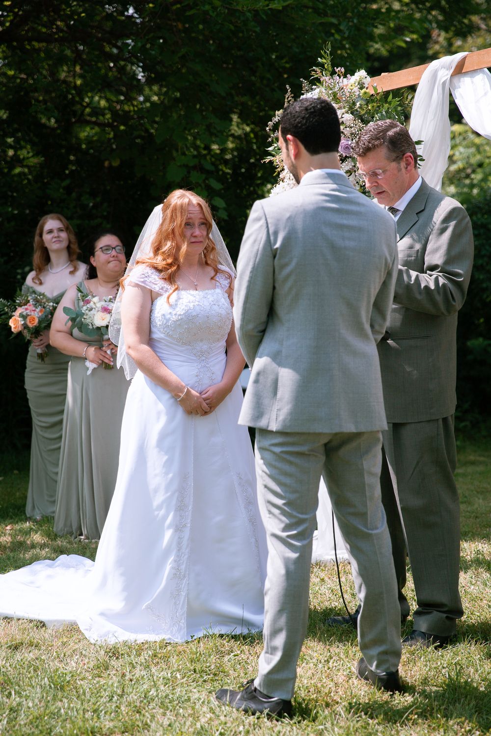 Bride in white lace gown smiling at groom during outdoor wedding ceremony with bridesmaids in sage green dresses.