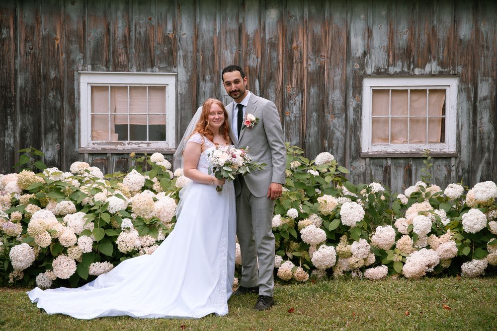 Smiling bride and groom posing together in front of rustic barn surrounded by white hydrangea bushes.