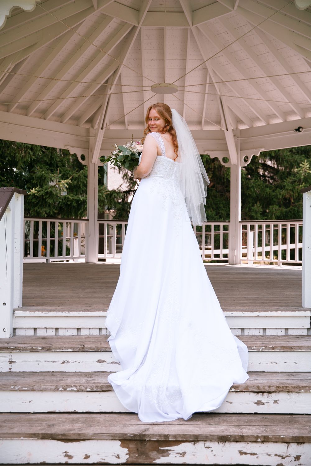 Bride in elegant white gown with veil holding bouquet, posing on steps of rustic white gazebo.