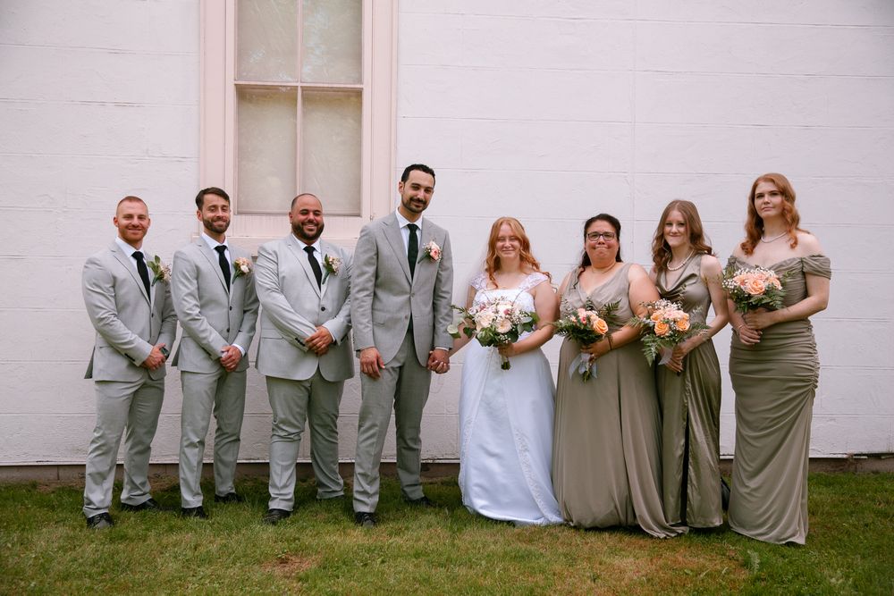Wedding party in gray suits and sage green dresses posing together outside white building.