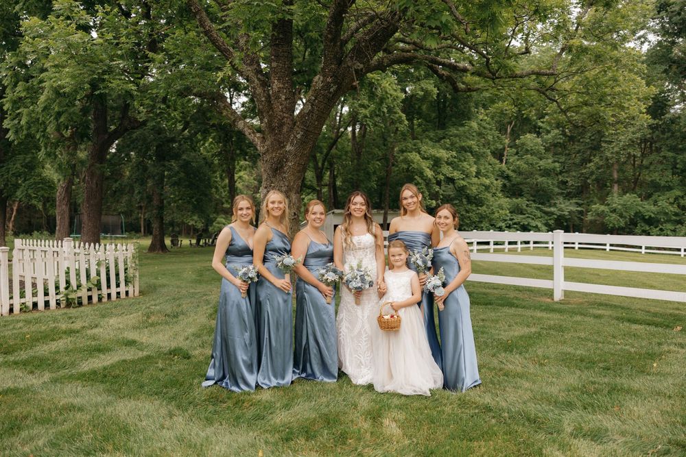 Wedding party in blue bridesmaid dresses poses on grassy lawn with white fences and trees in background.