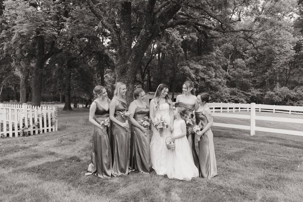 A group of people in formal attire pose for photos in a scenic outdoor garden setting with white fences and trees.
