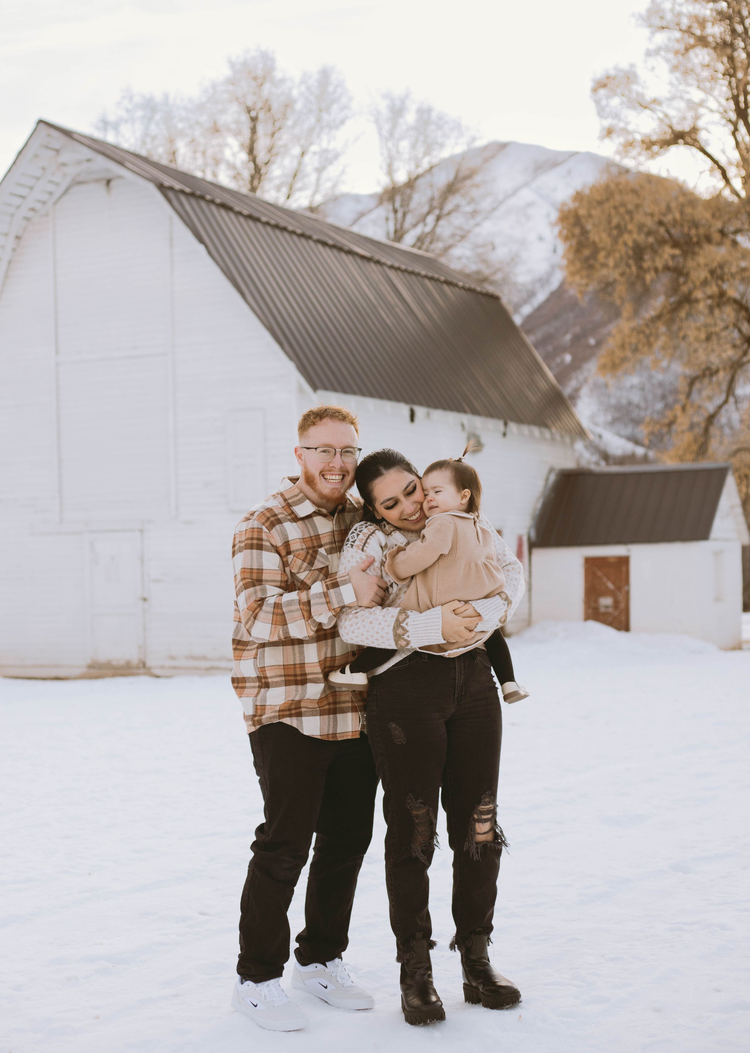 A loving couple embraces in a snowy winter setting with white barns and trees in the background.