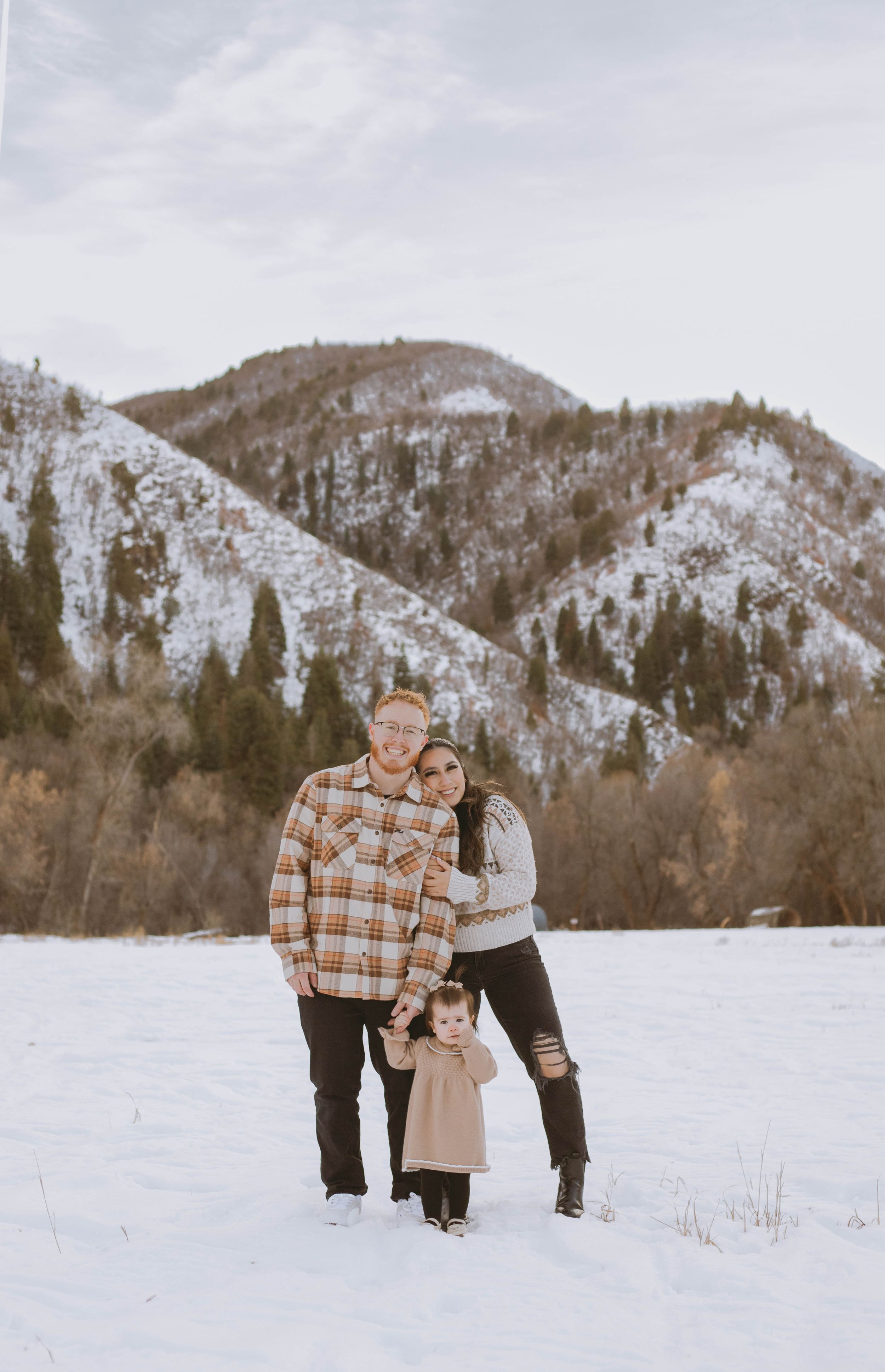 A family stands together in a snowy winter landscape with mountains and evergreen trees in the background.