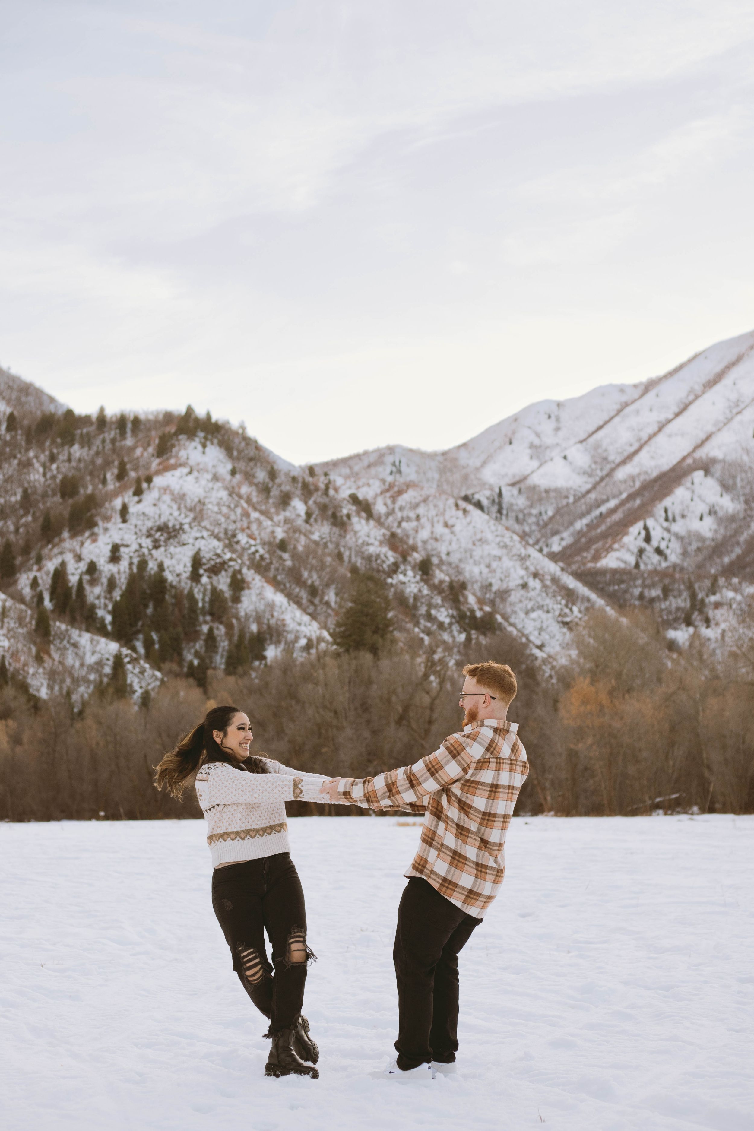 Two people holding hands and spinning in a snowy field with snow-covered mountains in the background on a winter day.