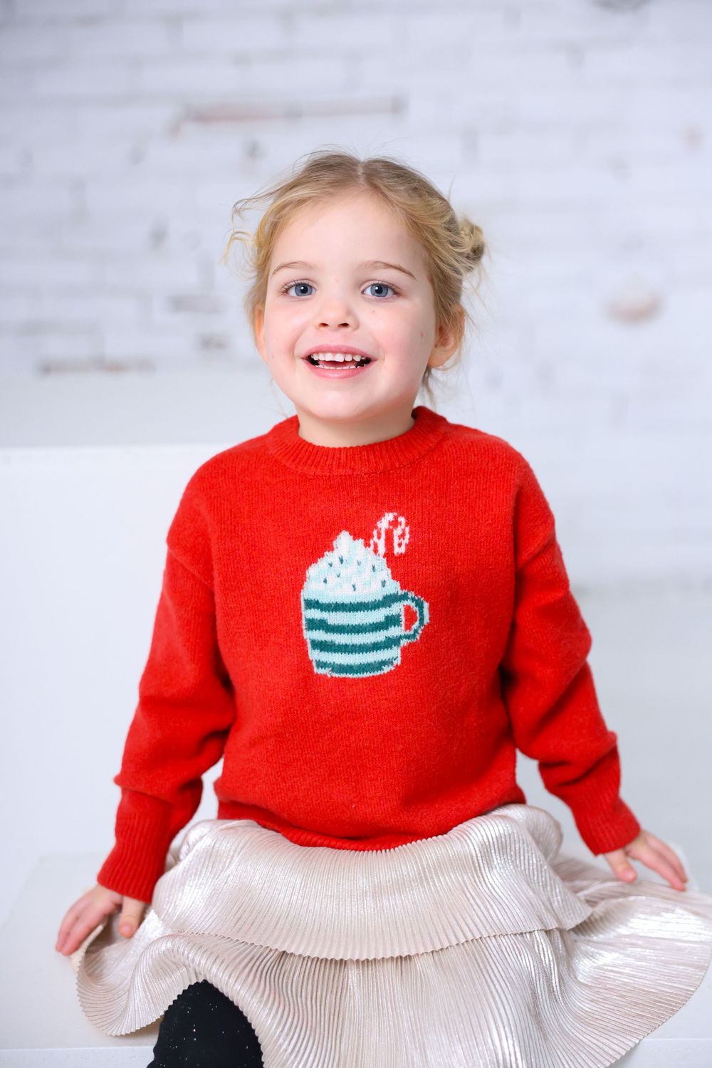 Child wearing festive red sweater with cute mug design against white brick background.