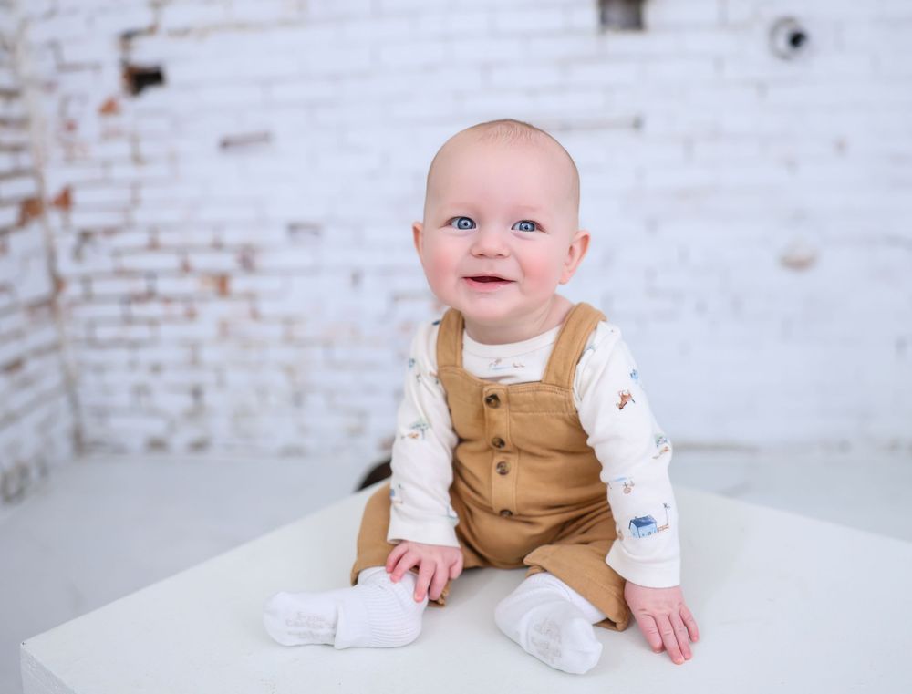 Baby in tan overalls sitting against white brick wall during studio photography session.