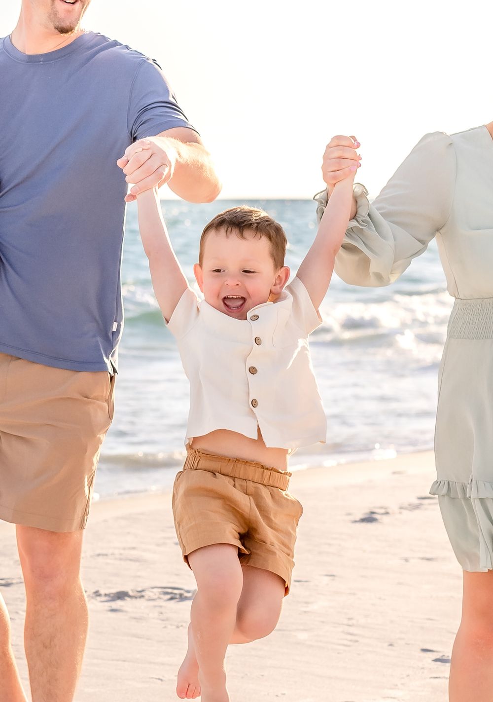 A joyful child in a white shirt and tan shorts jumps on the beach while holding hands with adults on a sunny day.