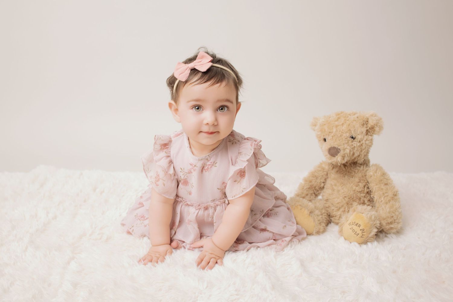 A baby in a pink dress sits with a teddy bear in a series of sweet studio portraits.