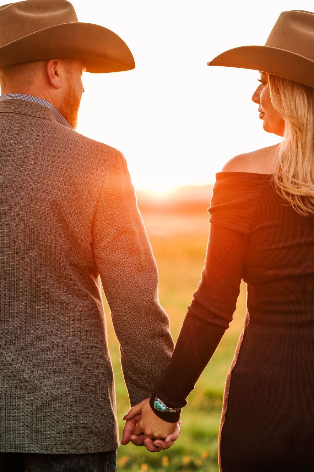 Silhouetted couple in cowboy hats hold hands against a warm sunset sky in a romantic countryside setting.