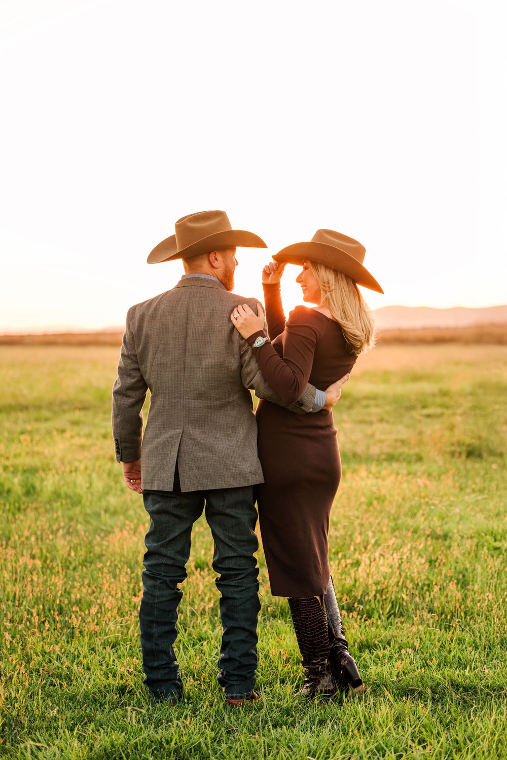 A couple wearing cowboy hats walks hand in hand through a golden field at sunset.