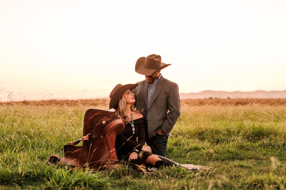 A cowboy rests in a grassy field beside a resting brown cow during sunset.