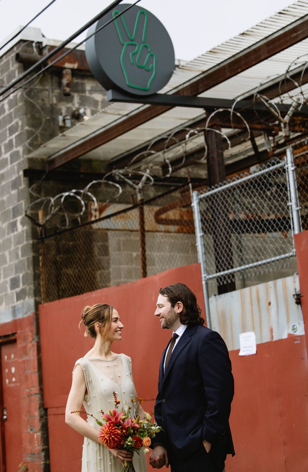 Couple's portrait at industrial-style venue space with red walls and chain-link fencing. Pizzeria Beddia Wedding. Fishtown Philadelphia, PA.