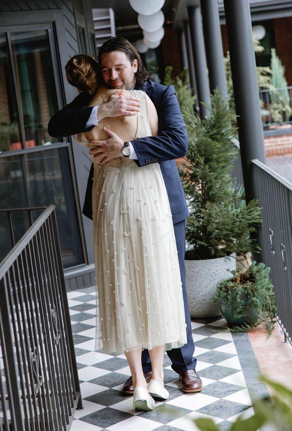A tender embrace captured on a porch with checkered flooring and potted greenery. Pizzeria Beddia Wedding. Fishtown Philadelphia, PA.