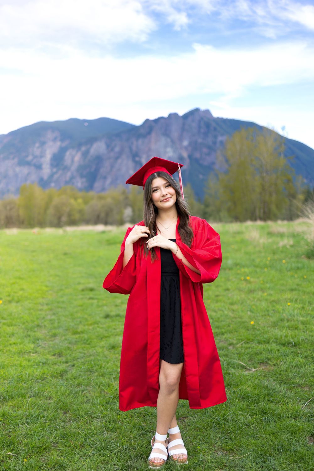 Young woman in red graduation cap and gown smiling outdoors with green fields and mountain backdrop.